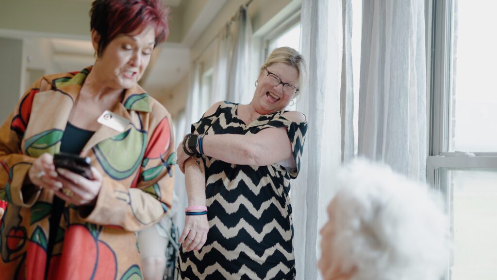 A woman sitting down is at a nursing home for a respite care visit and is greeted by a kind woman